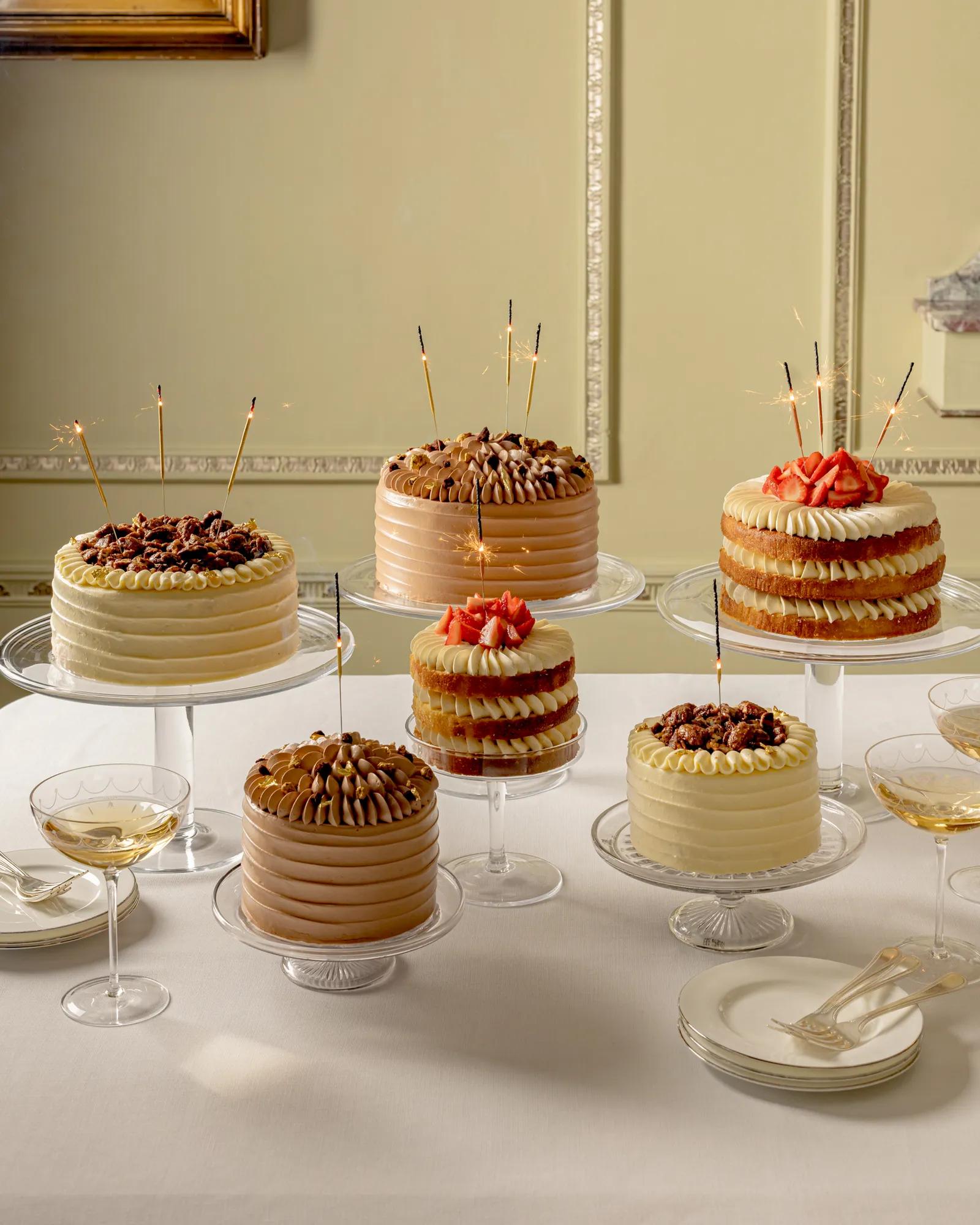 Celebration cakes on glass stands with sparklers, arranged on a table with dessert plates and coupe glasses.