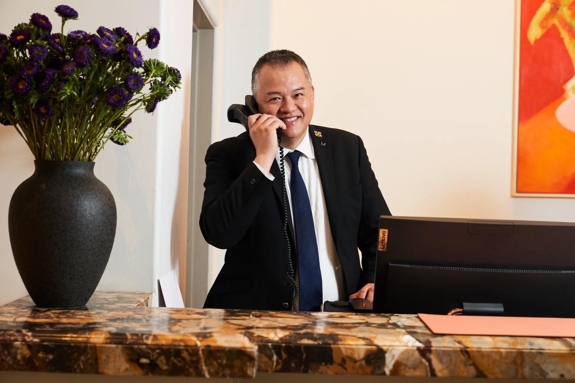 Concierge at The Maybourne Beverly Hills answering the phone behind the marble front desk with a vase of purple flowers.