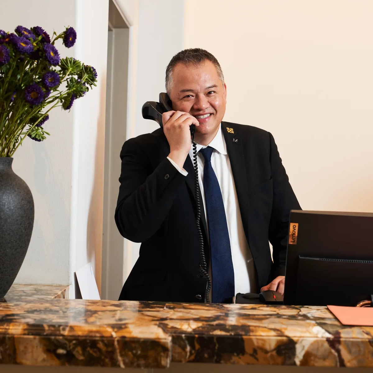 Concierge at The Maybourne Beverly Hills answering the phone behind the marble front desk with a vase of purple flowers.