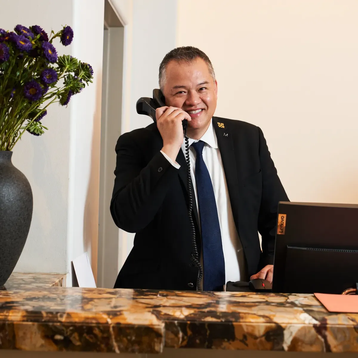 Concierge at The Maybourne Beverly Hills answering the phone behind the marble front desk with a vase of purple flowers.