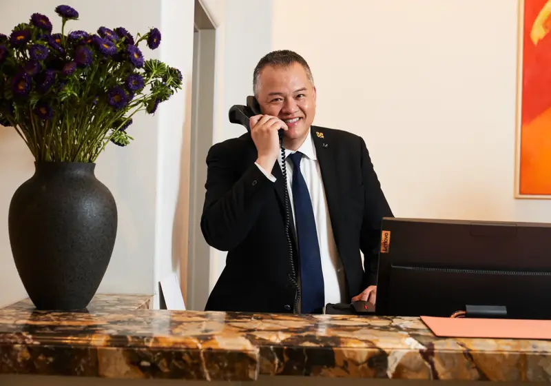 Concierge at The Maybourne Beverly Hills answering the phone behind the marble front desk with a vase of purple flowers.
