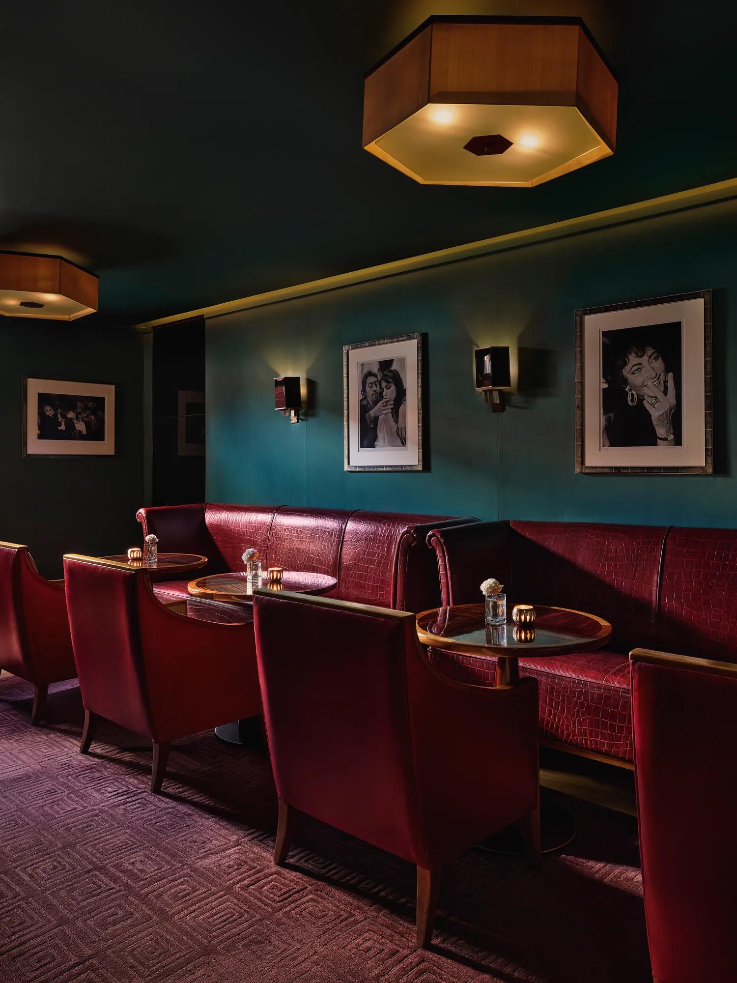 Intimate lounge area with red leather armchairs and banquette seating around small circular tables, teal walls decorated with black-and-white portraits, and warm ambient lighting.