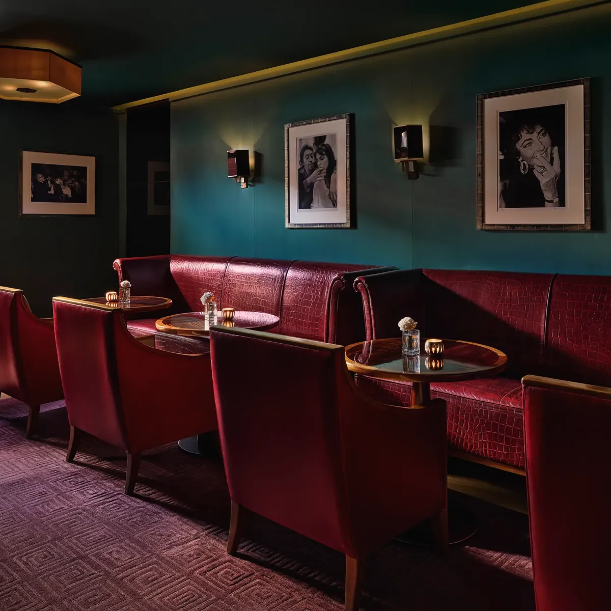 Intimate lounge area with red leather armchairs and banquette seating around small circular tables, teal walls decorated with black-and-white portraits, and warm ambient lighting.