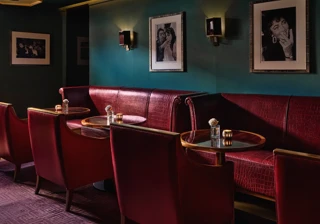 Intimate lounge area with red leather armchairs and banquette seating around small circular tables, teal walls decorated with black-and-white portraits, and warm ambient lighting.