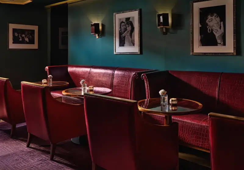 Intimate lounge area with red leather armchairs and banquette seating around small circular tables, teal walls decorated with black-and-white portraits, and warm ambient lighting.