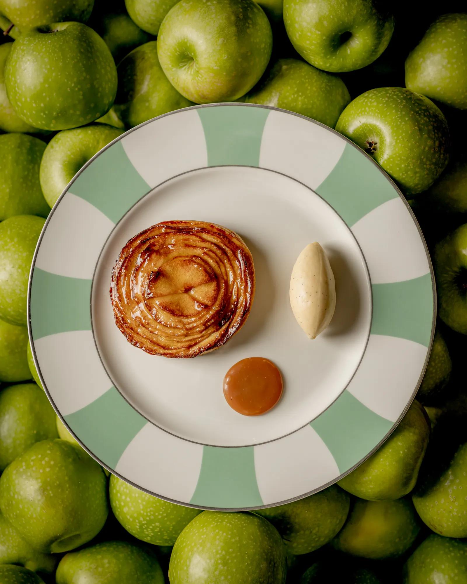 Apple tart with quenelle of ice cream and caramel sauce on striped plate surrounded by green apples.