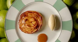 Apple tart with quenelle of ice cream and caramel sauce on striped plate surrounded by green apples.