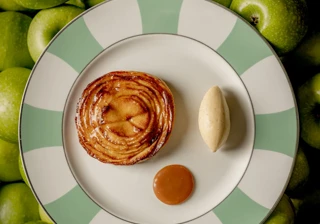 Apple tart with quenelle of ice cream and caramel sauce on striped plate surrounded by green apples.