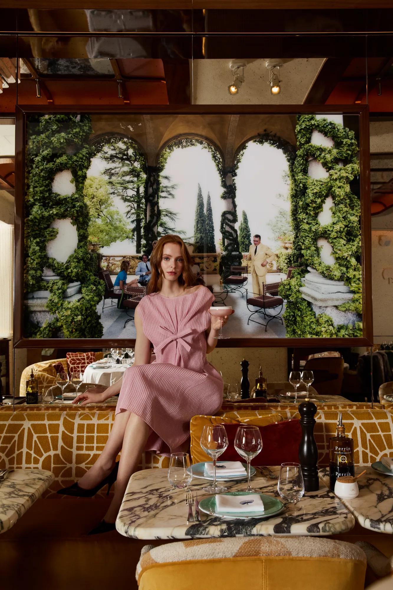 A person in a pink knotted dress sits on a patterned restaurant banquette holding a cocktail glass. In the foreground, marble-topped tables are set with plates, wine glasses, and a pepper mill. Behind the seating area, a large mural or window-like feature framed by lush green foliage depicts an outdoor terrace with arches, trees, and café seating, creating an elegant, Mediterranean-inspired atmosphere.