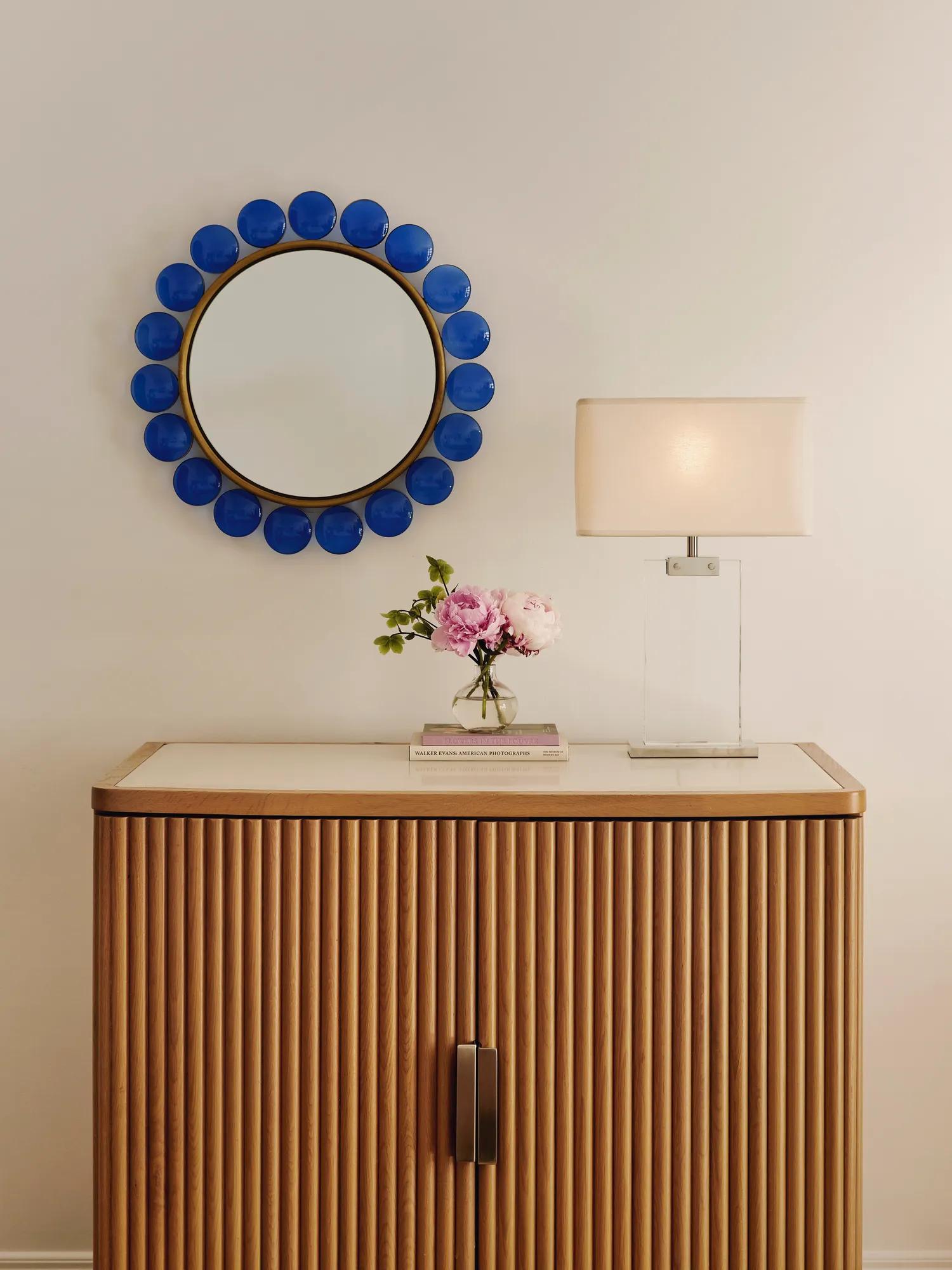 Wooden cabinet with lamp, flowers, and books beneath a round mirror framed with bold blue accents.