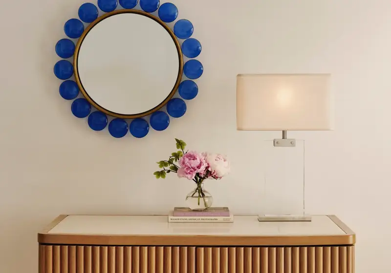 Wooden cabinet with lamp, flowers, and books beneath a round mirror framed with bold blue accents.