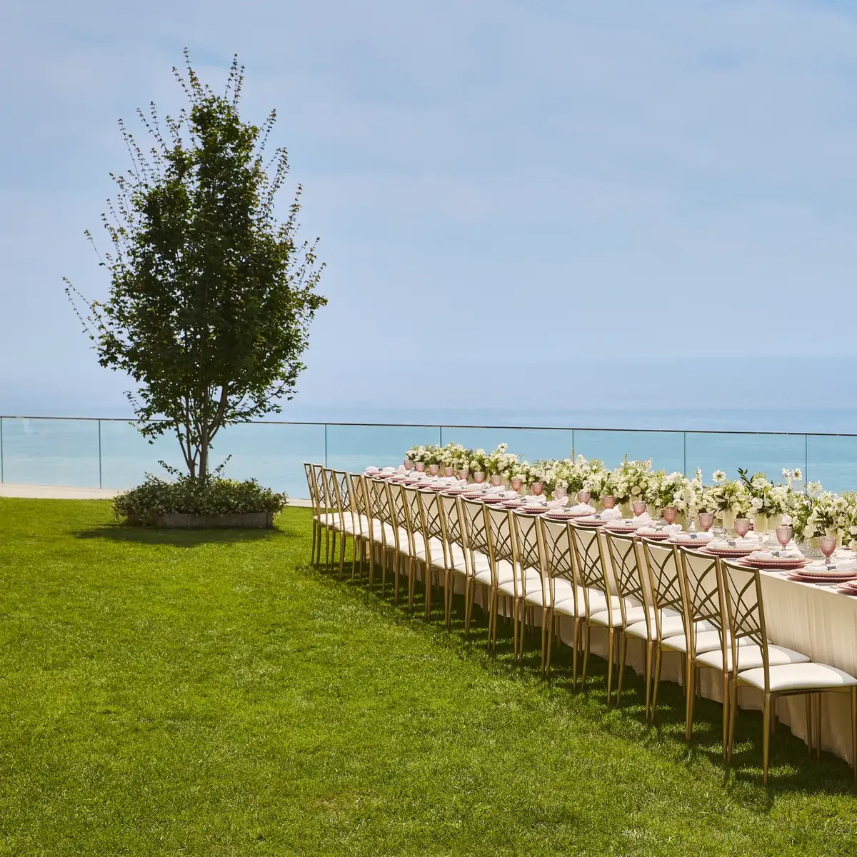 Élégante table de banquet dressée avec des arrangements floraux blancs et des assiettes roses sur une pelouse surplombant la mer.