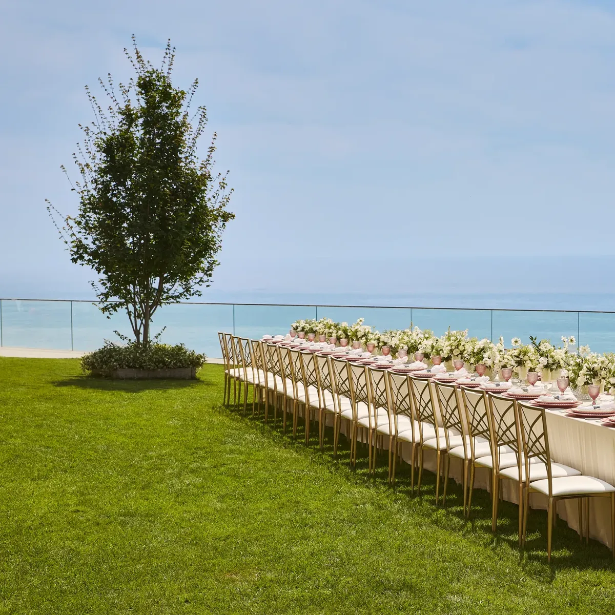 Élégante table de banquet dressée avec des arrangements floraux blancs et des assiettes roses sur une pelouse surplombant la mer.