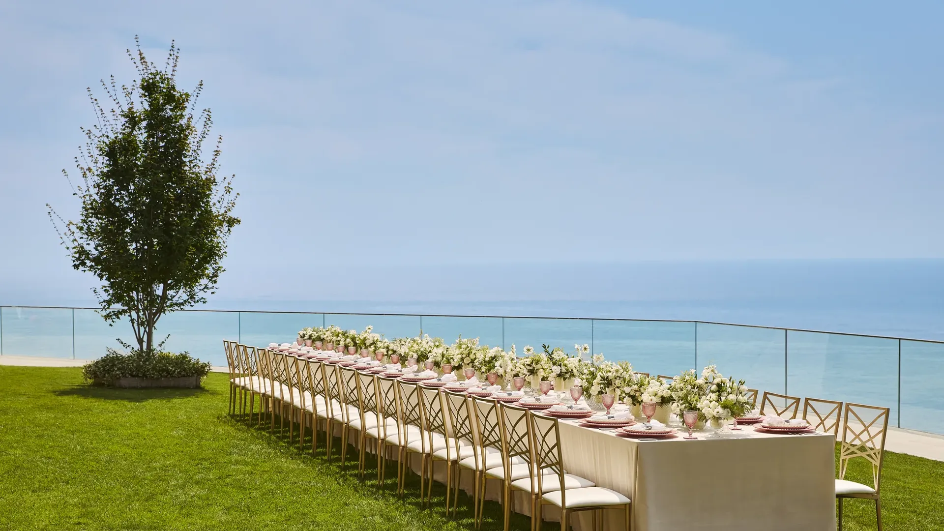 Élégante table de banquet dressée avec des arrangements floraux blancs et des assiettes roses sur une pelouse surplombant la mer.
