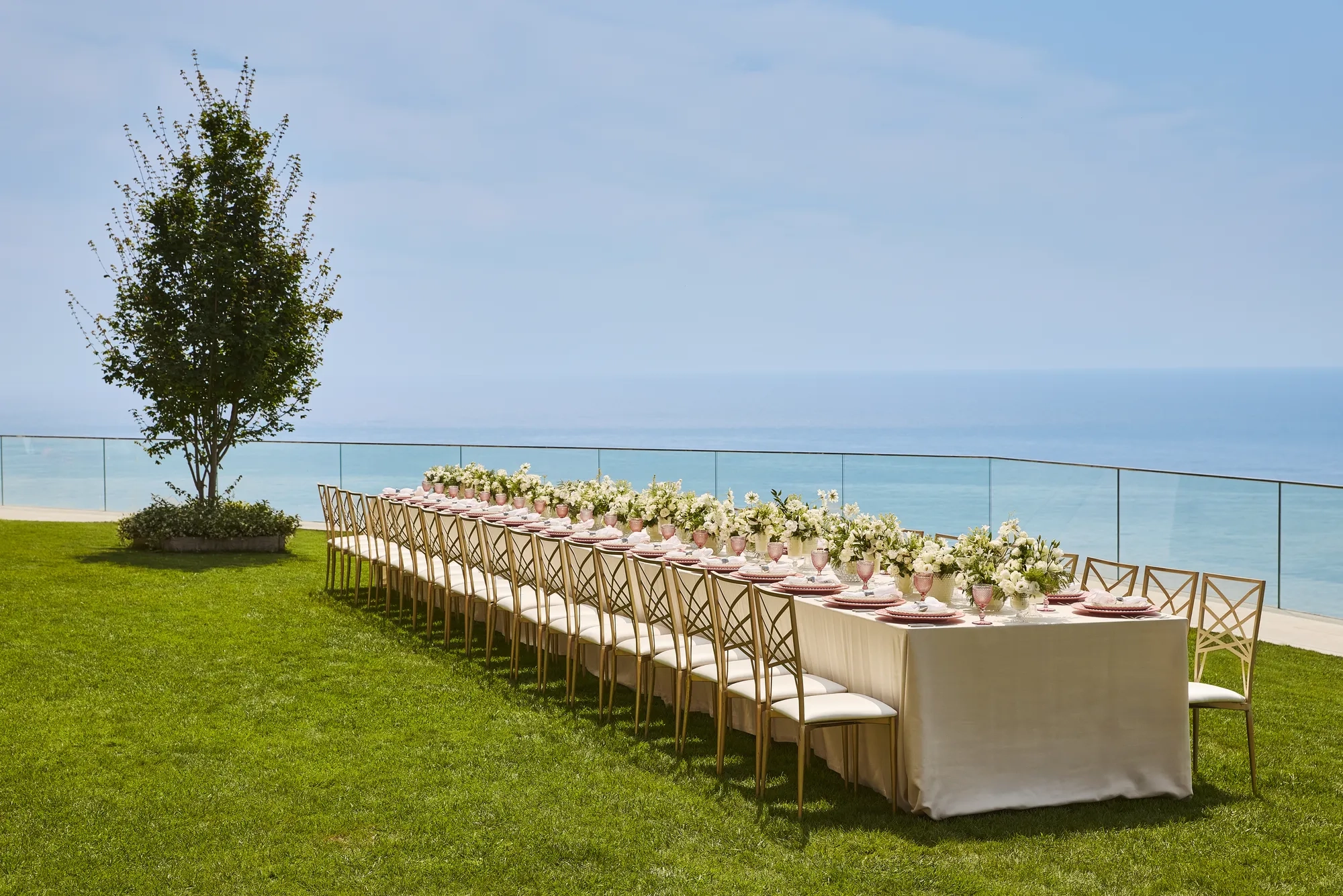 Élégante table de banquet dressée avec des arrangements floraux blancs et des assiettes roses sur une pelouse surplombant la mer.