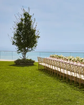 Élégante table de banquet dressée avec des arrangements floraux blancs et des assiettes roses sur une pelouse surplombant la mer.