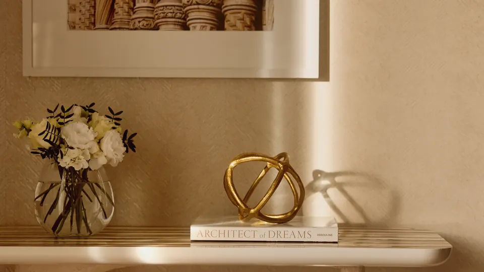 Close-up of a cream console table with a vase of flowers, decorative gold sculpture, and a book beneath a framed photograph of architectural column