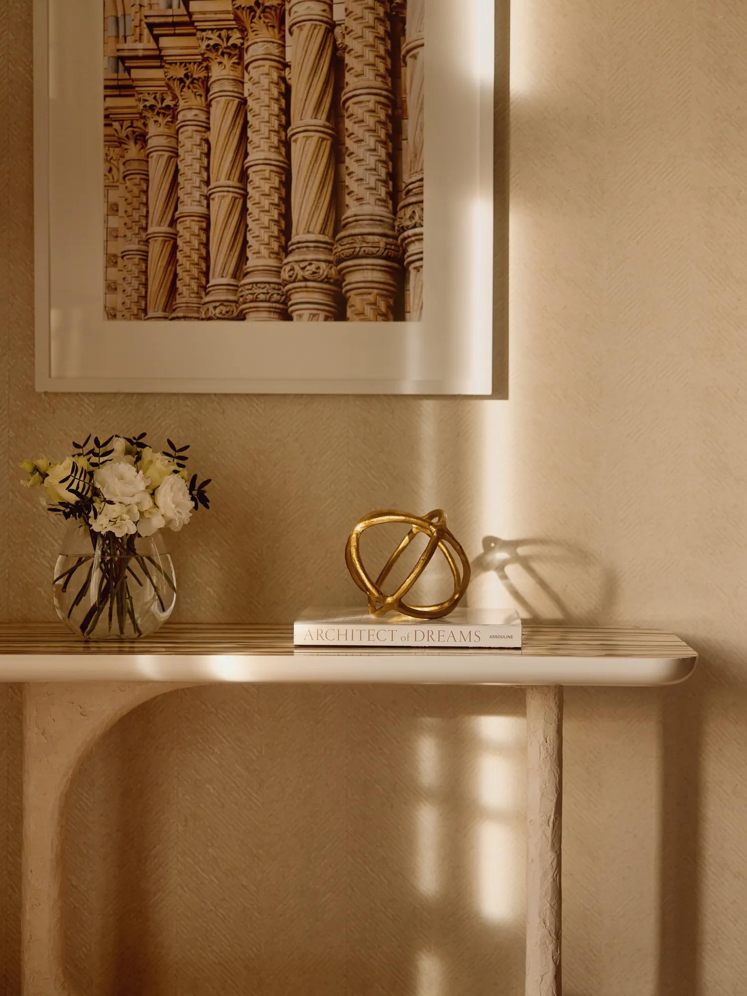 Close-up of a cream console table with a vase of flowers, decorative gold sculpture, and a book beneath a framed photograph of architectural column