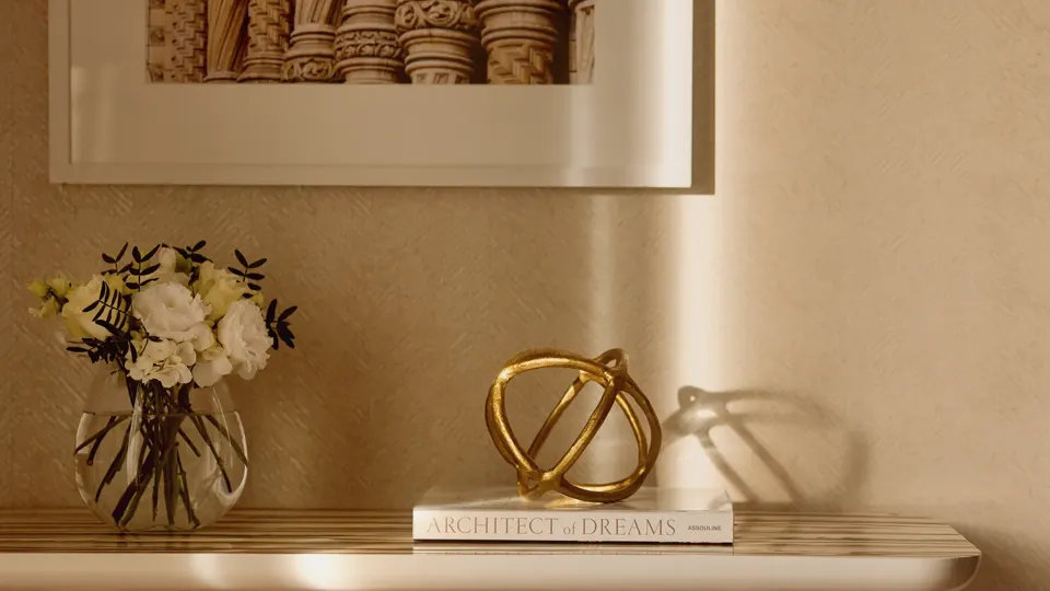 Close-up of a cream console table with a vase of flowers, decorative gold sculpture, and a book beneath a framed photograph of architectural column