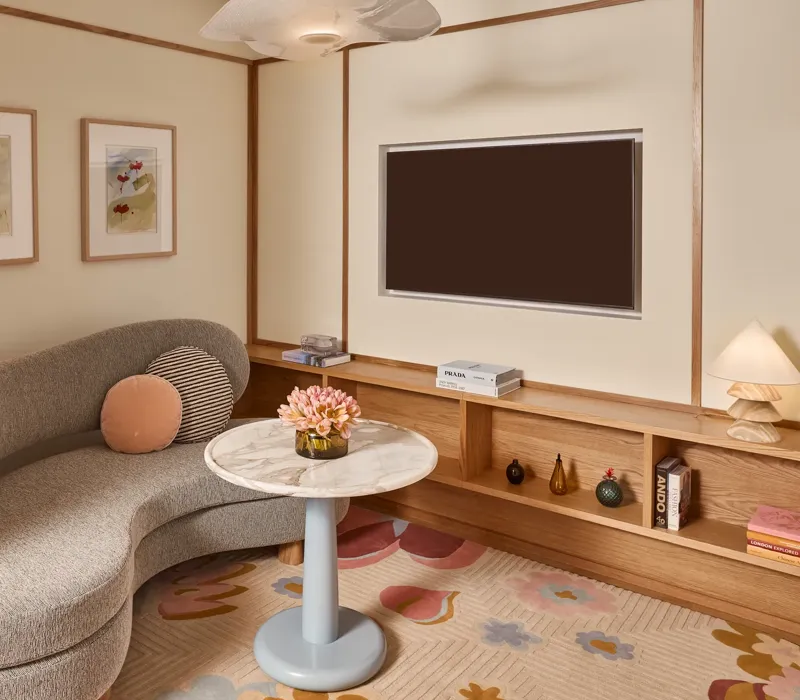 Cosy sitting area with curved sofa, marble-topped table, wall-mounted TV, built-in wooden shelving, and framed artwork.