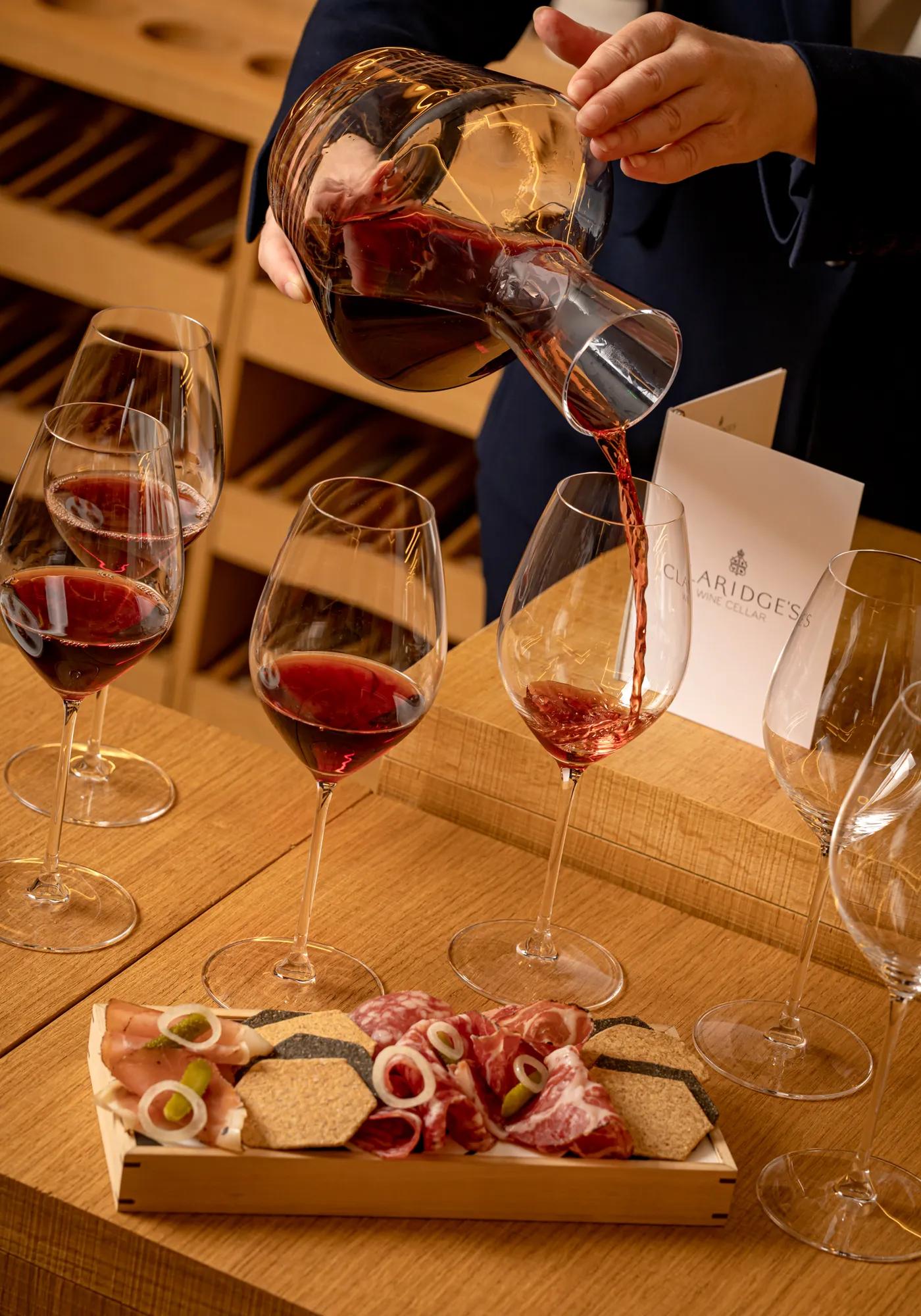 A decanter pours red wine into glasses on a wooden tasting table, with a charcuterie board and Claridge’s Wine Cellar menu in view.