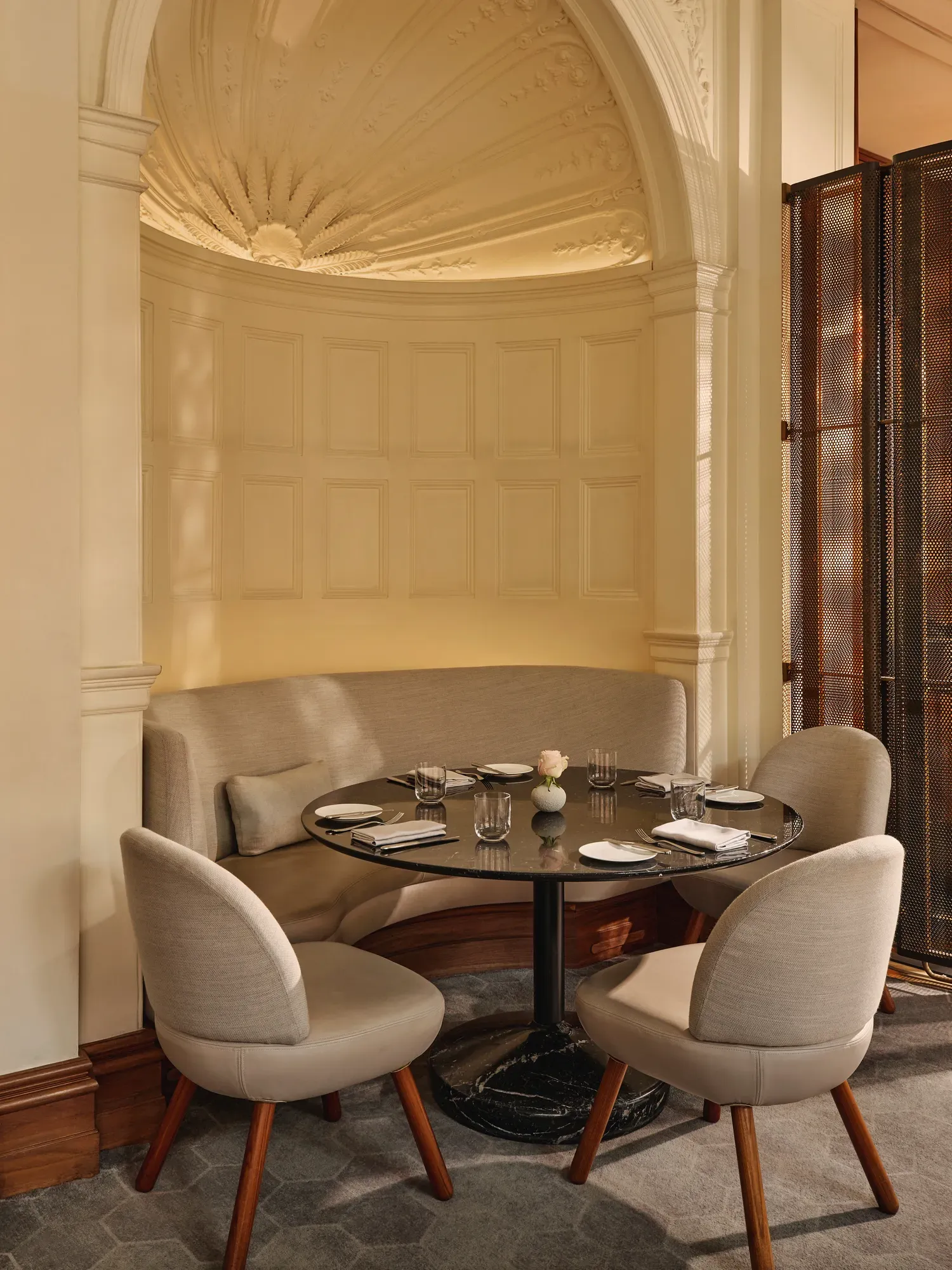 Small dining nook with a curved upholstered banquette, round black table, and soft beige chairs beneath an ornate domed ceiling.