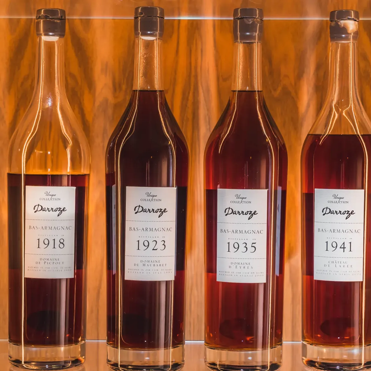 Four vintage Darroze Bas-Armagnac bottles with dated labels displayed against a warm wooden backdrop on glass shelf