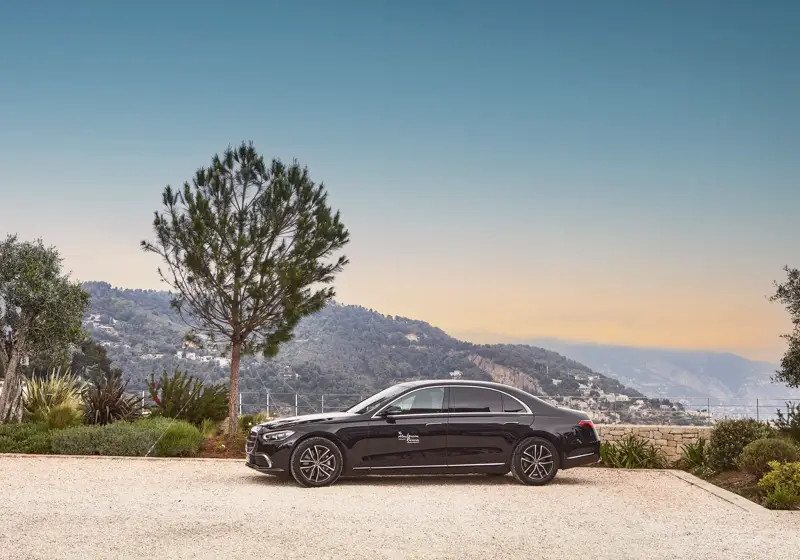Black luxury hotel car parked overlooking hillside towns and a hazy coastline at sunset.