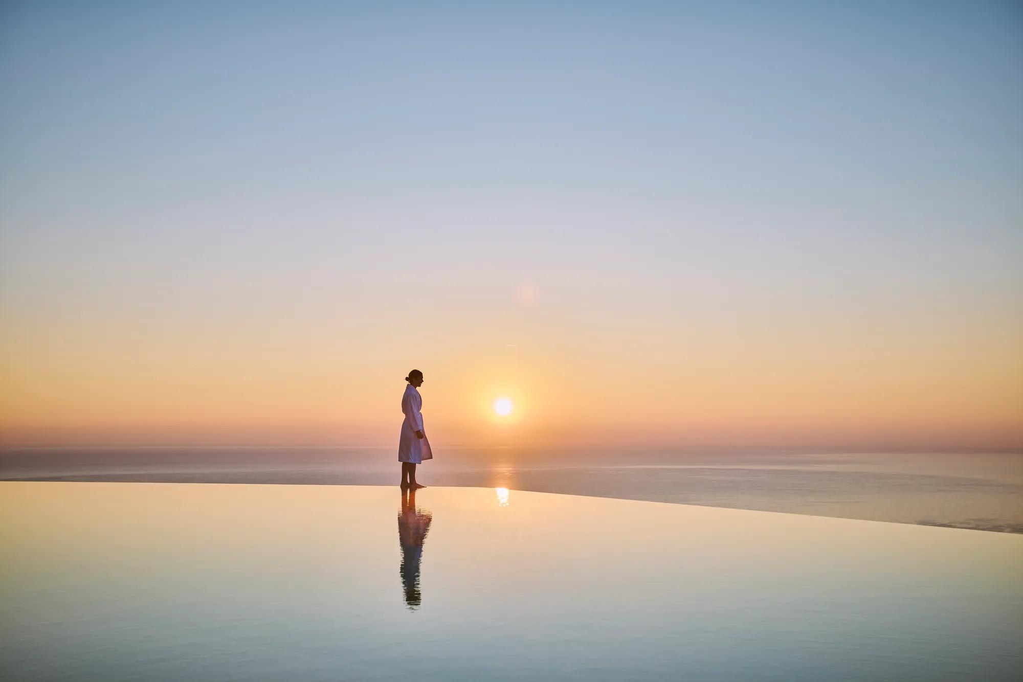 Silhouetted figure standing beside an infinity pool at sunrise, overlooking a calm sea and horizon.
