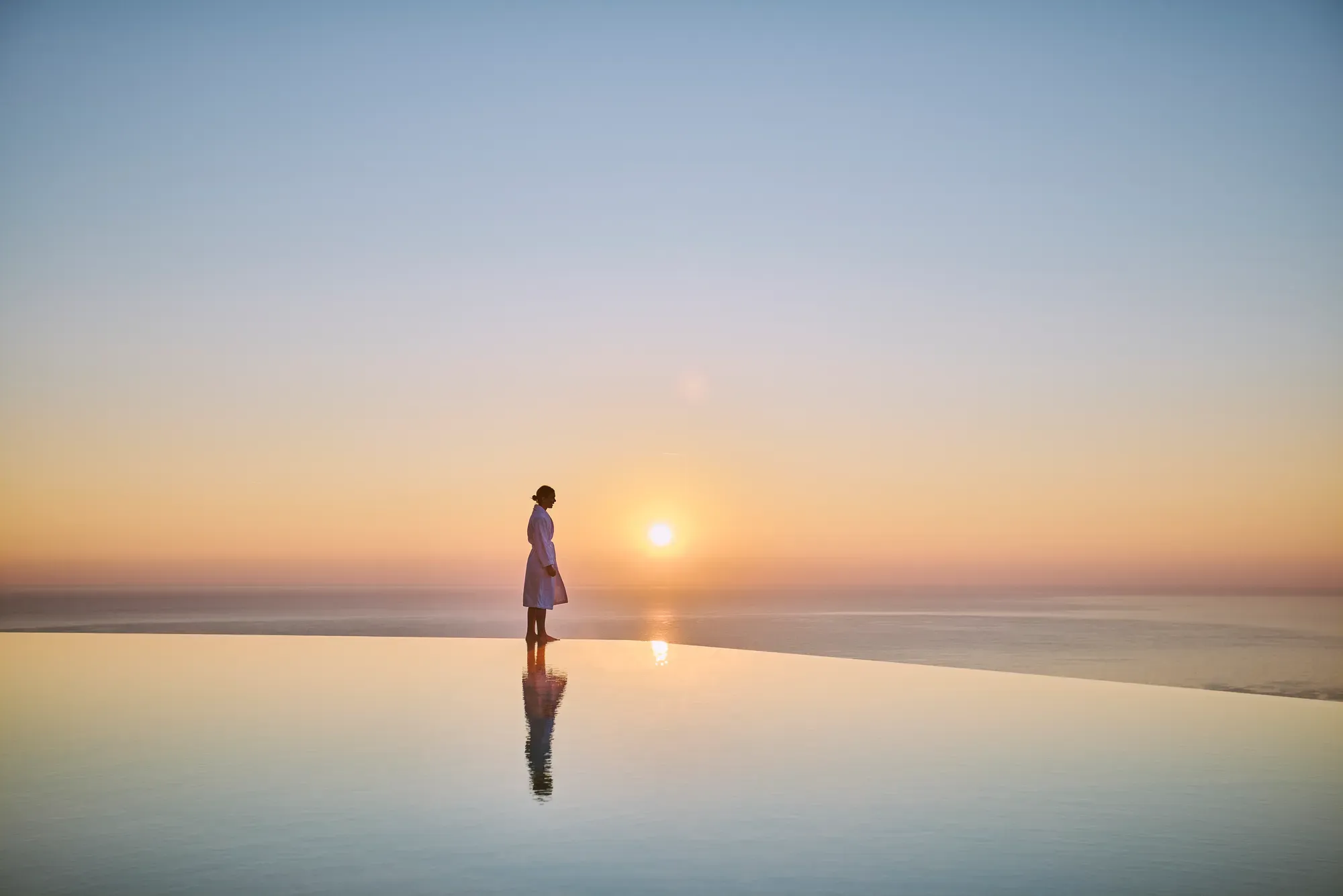 Silhouetted figure standing beside an infinity pool at sunrise, overlooking a calm sea and horizon.