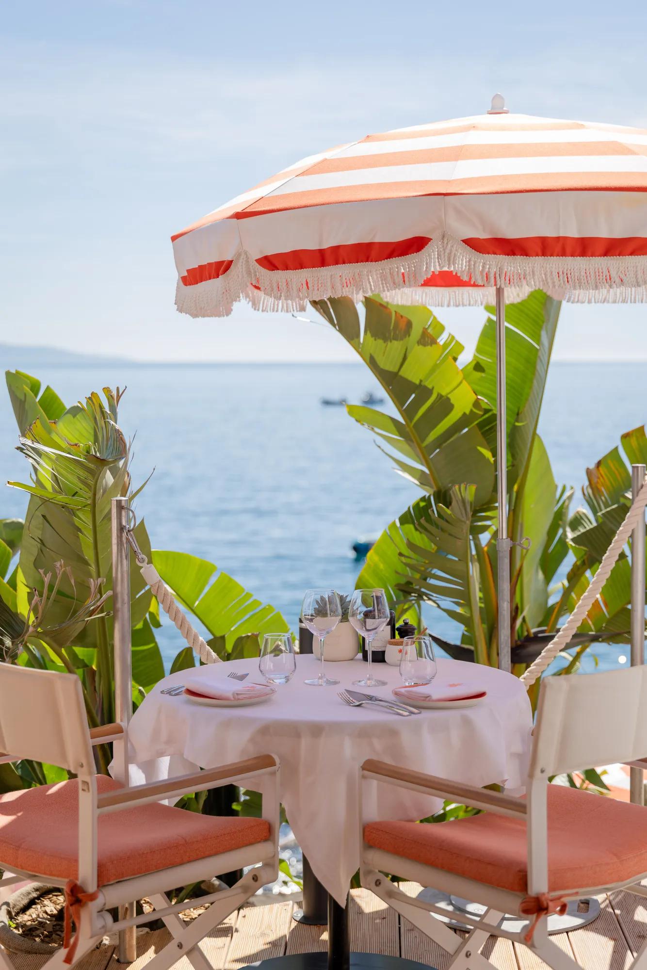 Table pour deux en bord de mer sous un parasol rayé, avec chaises corail et vue sur la mer.