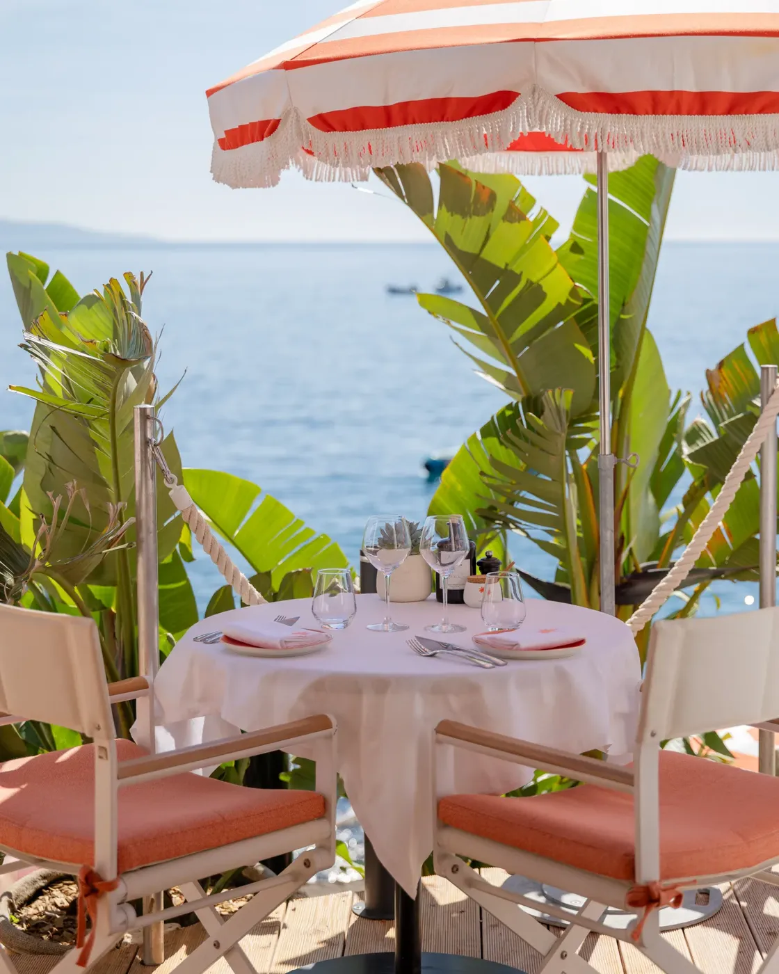 Table pour deux en bord de mer sous un parasol rayé, avec chaises corail et vue sur la mer.