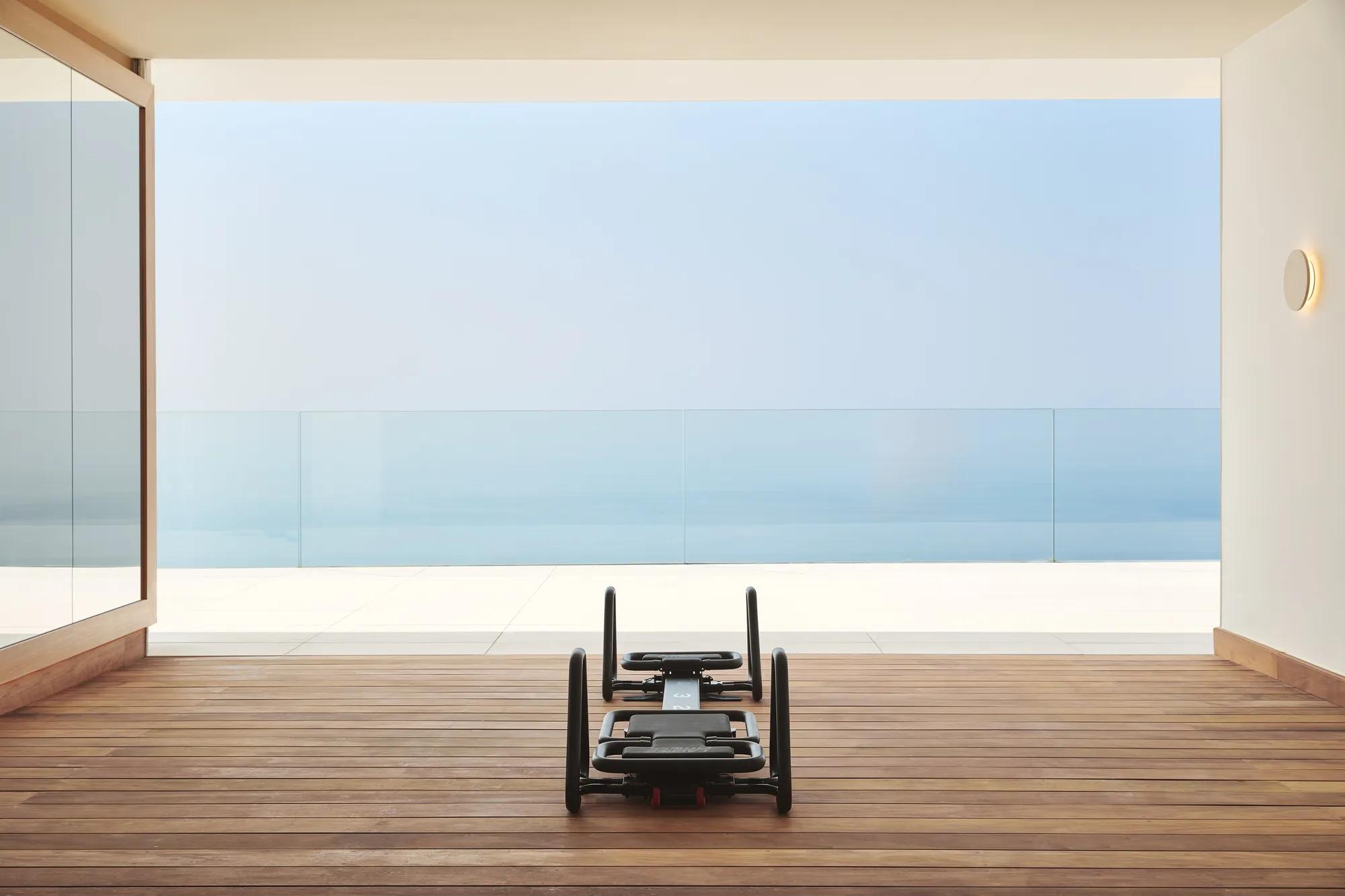 Pilates reformer on a wooden terrace overlooking the sea through floor-to-ceiling glass.