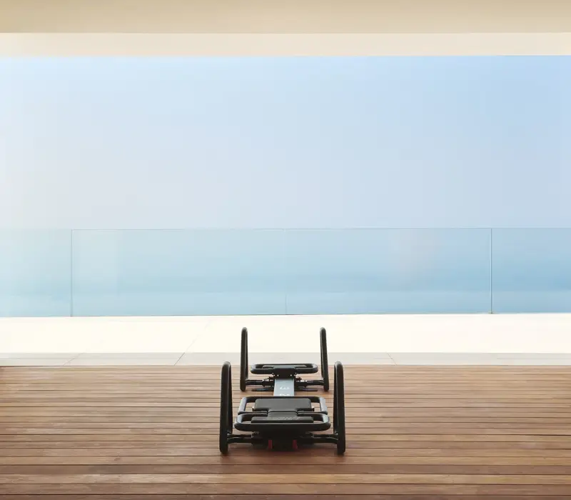 Pilates reformer on a wooden terrace overlooking the sea through floor-to-ceiling glass.