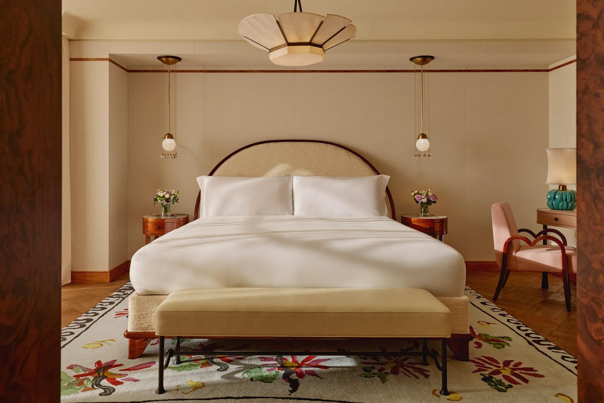 Inviting bedroom with a king-size bed dressed in white linens, arched headboard, pendant bedside lights, and floral-patterned rug, complemented by a pink armchair and turquoise lamp on a side table.