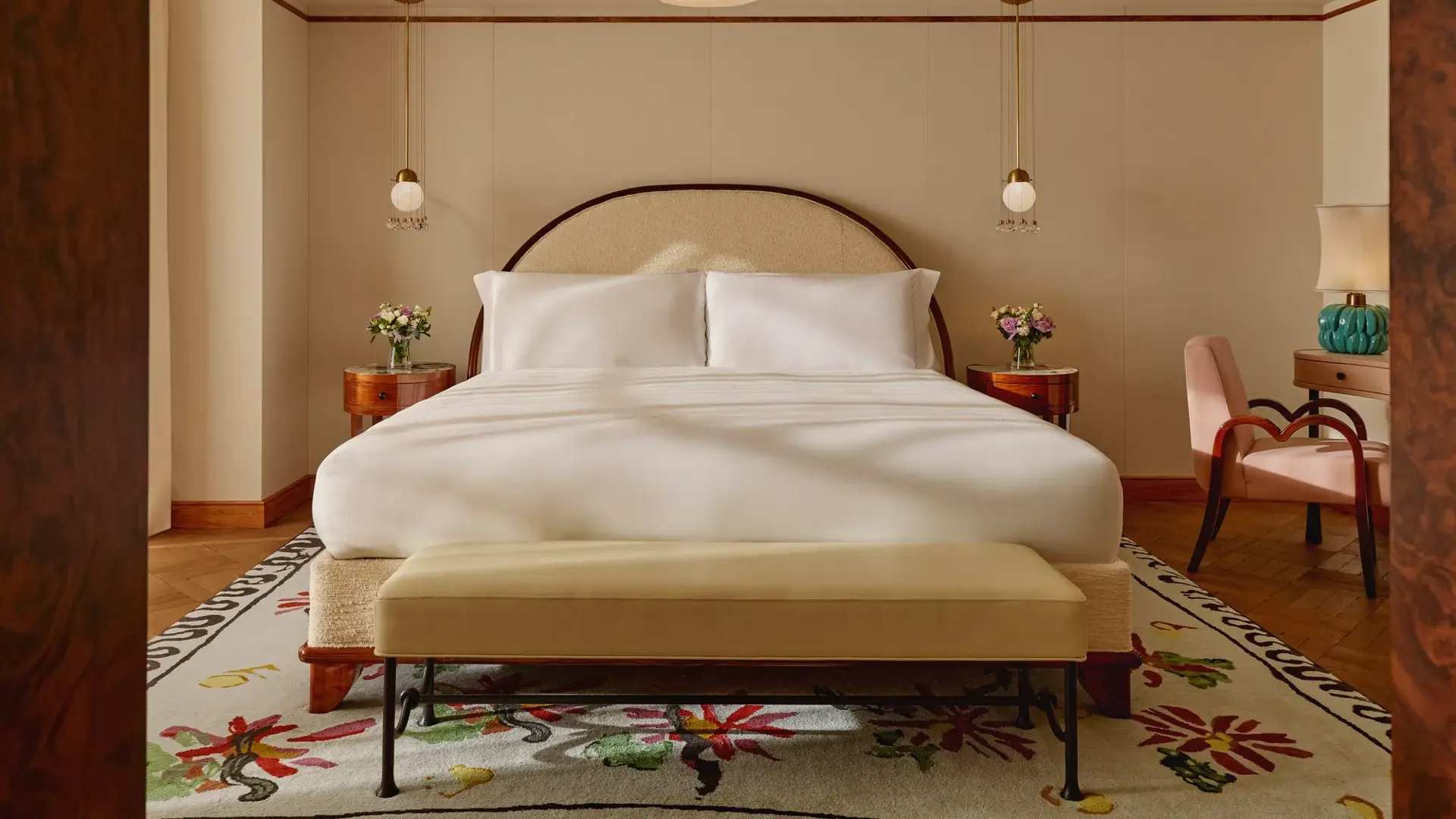 Inviting bedroom with a king-size bed dressed in white linens, arched headboard, pendant bedside lights, and floral-patterned rug, complemented by a pink armchair and turquoise lamp on a side table.
