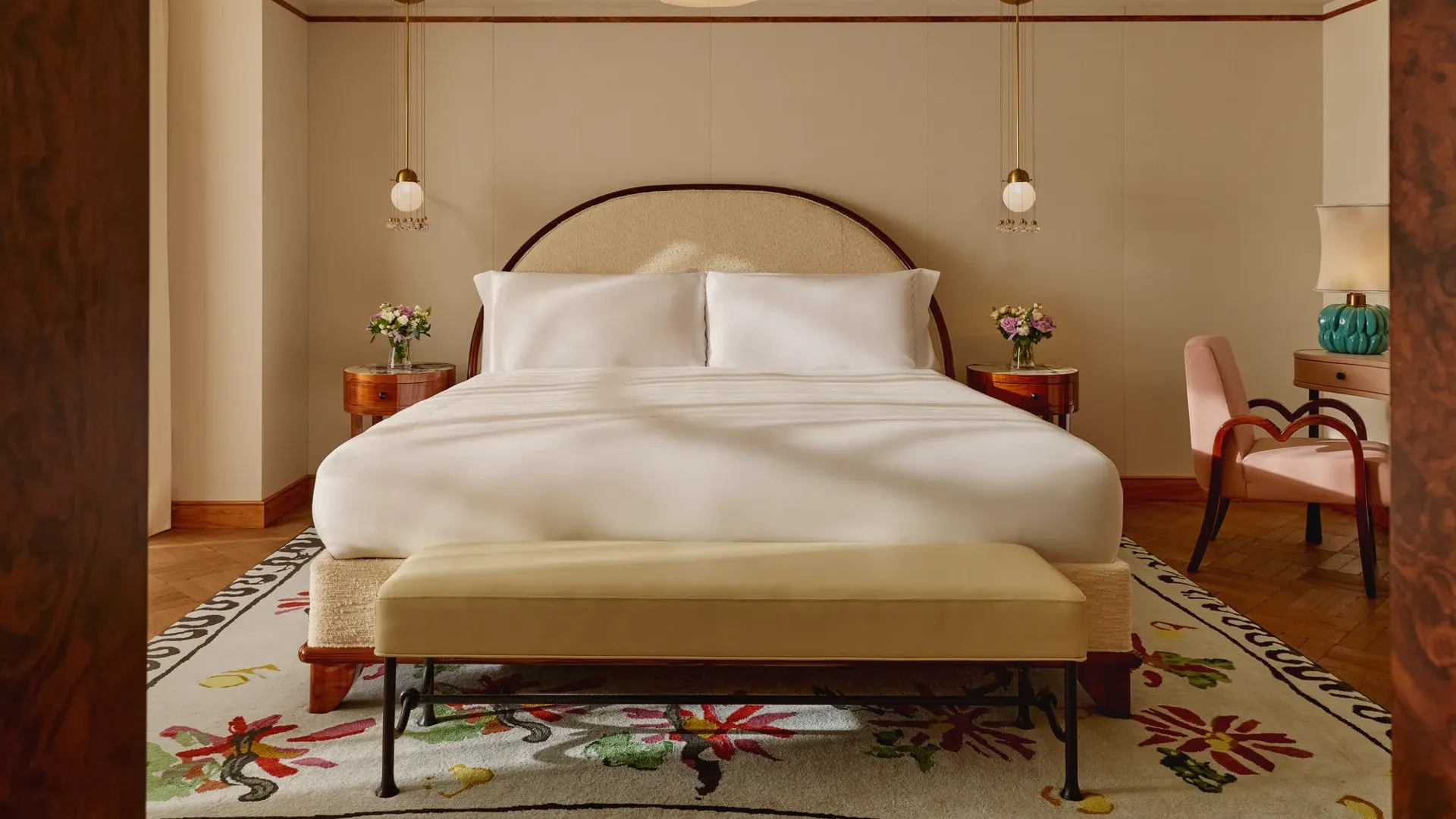 Inviting bedroom with a king-size bed dressed in white linens, arched headboard, pendant bedside lights, and floral-patterned rug, complemented by a pink armchair and turquoise lamp on a side table.