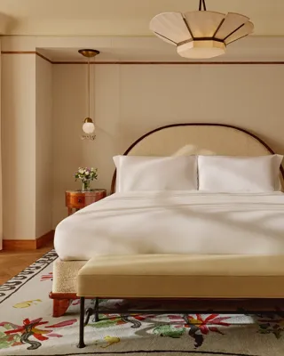 Inviting bedroom with a king-size bed dressed in white linens, arched headboard, pendant bedside lights, and floral-patterned rug, complemented by a pink armchair and turquoise lamp on a side table.