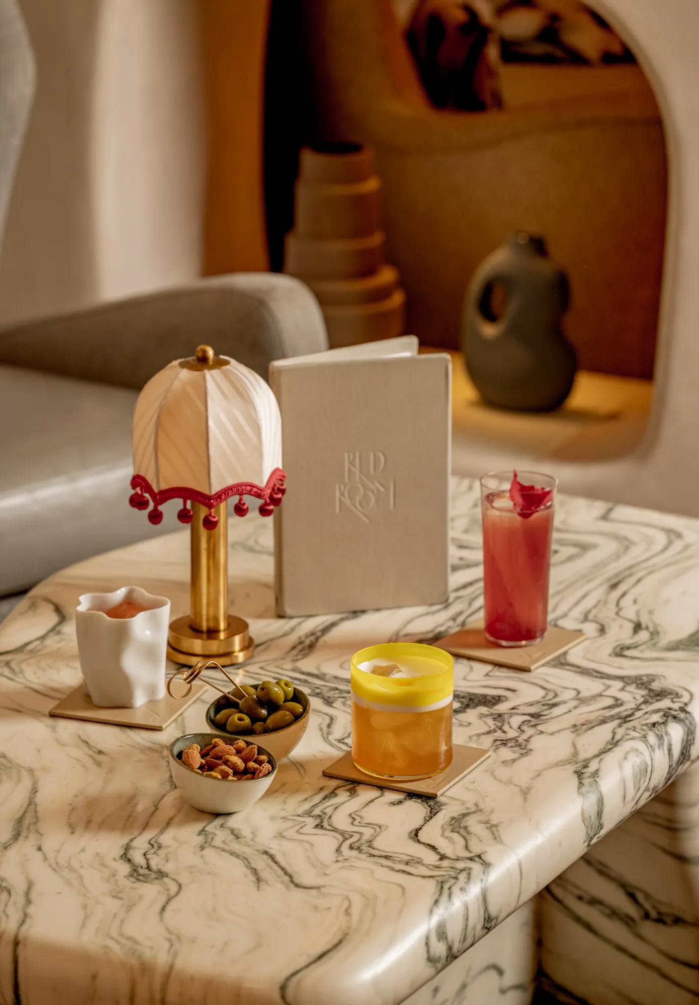 Marble coffee table styled with a pink cocktail, olives, nuts, candle, sculptural lamp and menu in a warm, contemporary lounge.