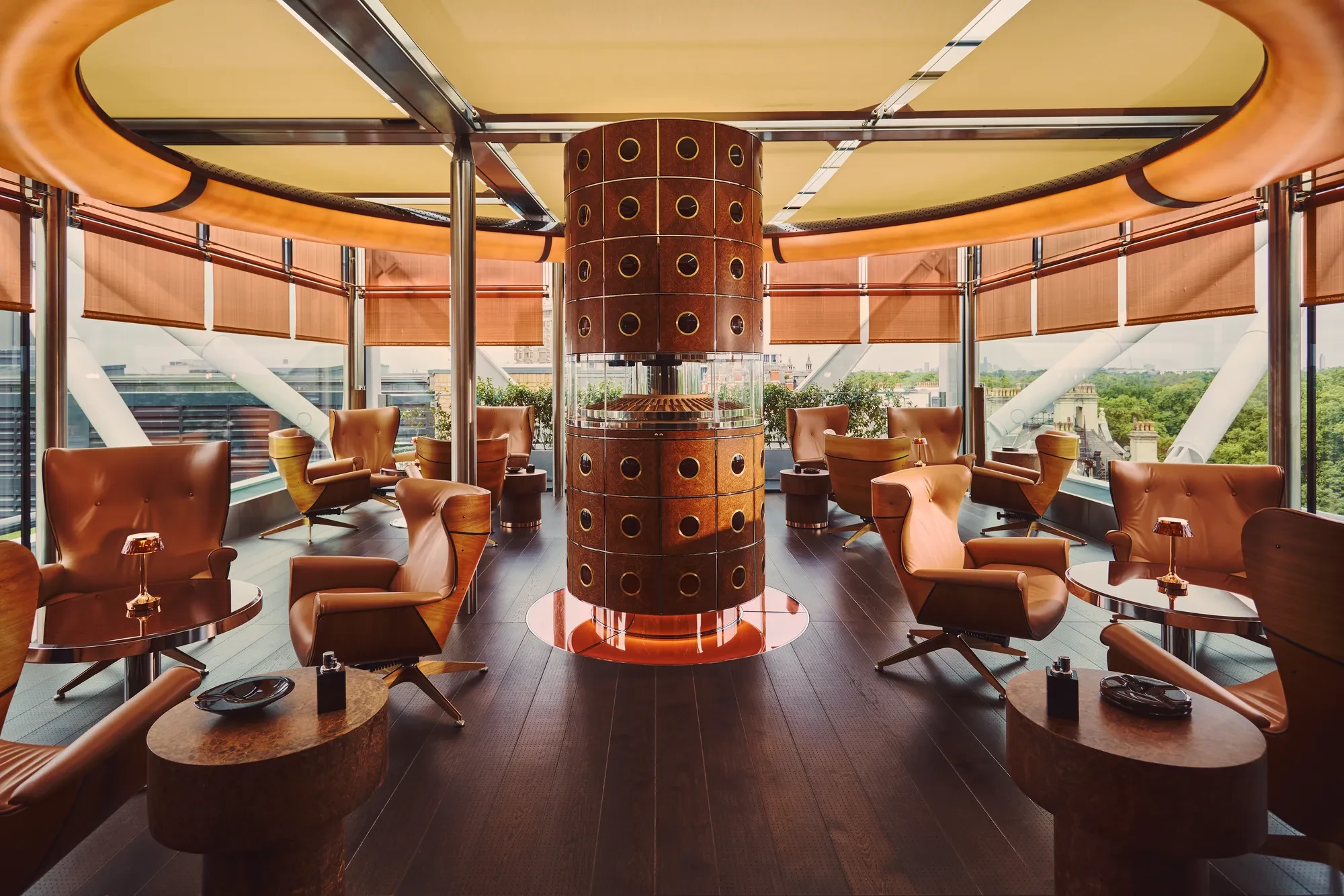 Rooftop lounge with tan leather chairs around small tables, a sculptural copper column centrepiece, and panoramic city views through glass walls.