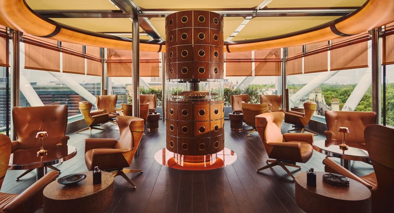 Rooftop lounge with tan leather chairs around small tables, a sculptural copper column centrepiece, and panoramic city views through glass walls.