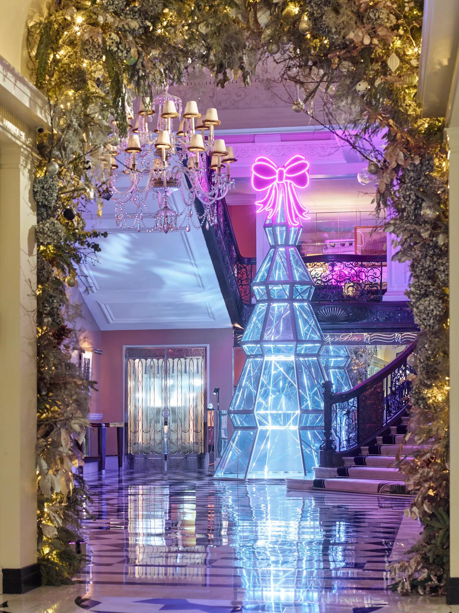 Festive hotel lobby with illuminated geometric tree sculpture and neon bow, framed by greenery and chandeliers, reflected on polished checkered floor.