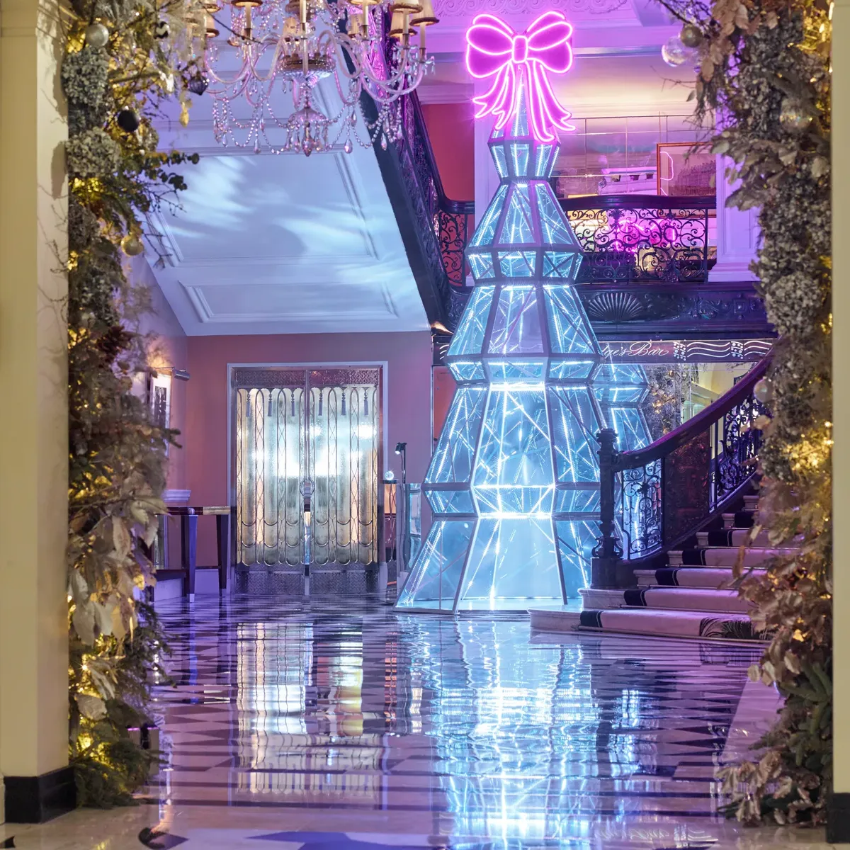 Festive hotel lobby with illuminated geometric tree sculpture and neon bow, framed by greenery and chandeliers, reflected on polished checkered floor.