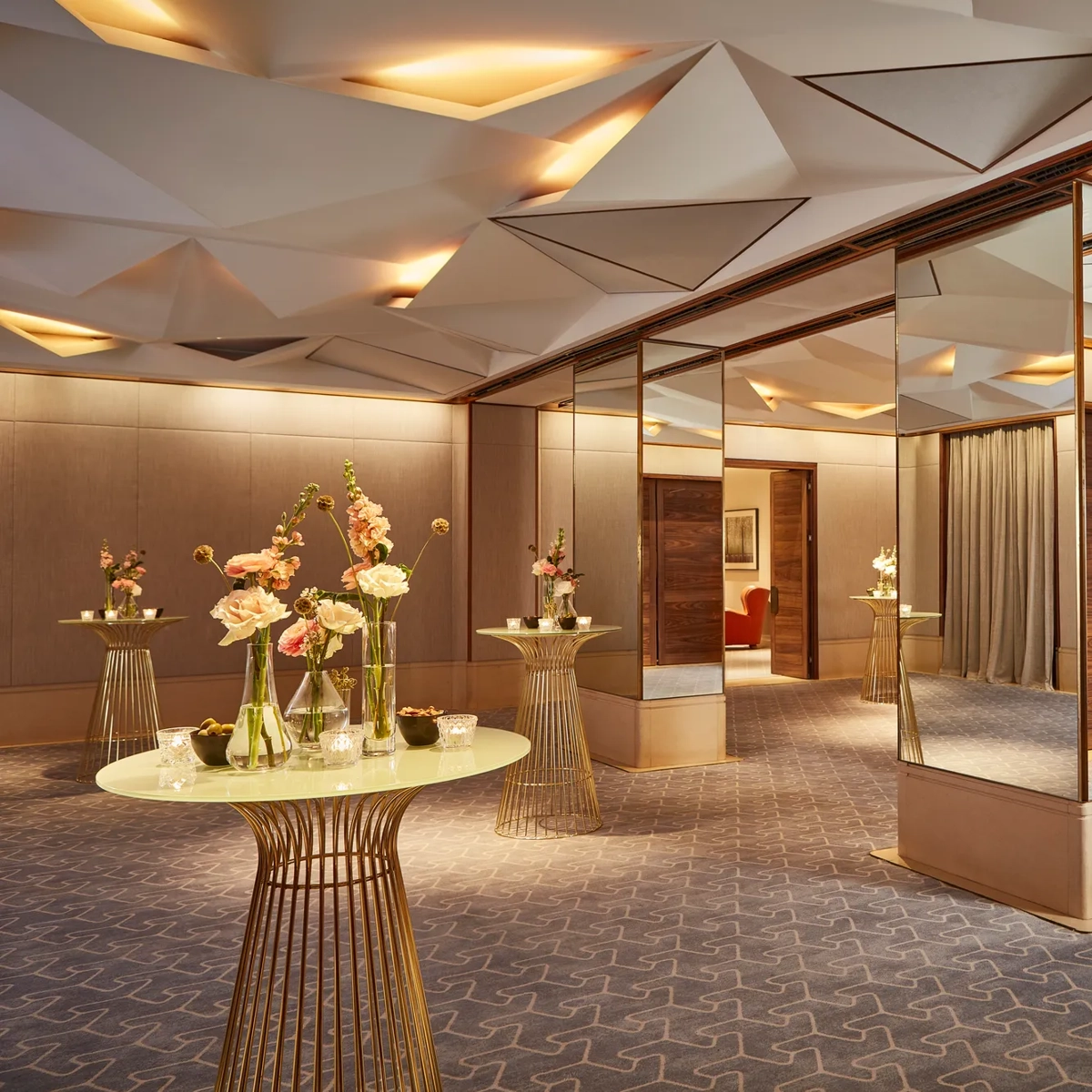 Elegant pre-function space with mirrored columns, gold tables, and floral decorations under geometric ceiling lights.