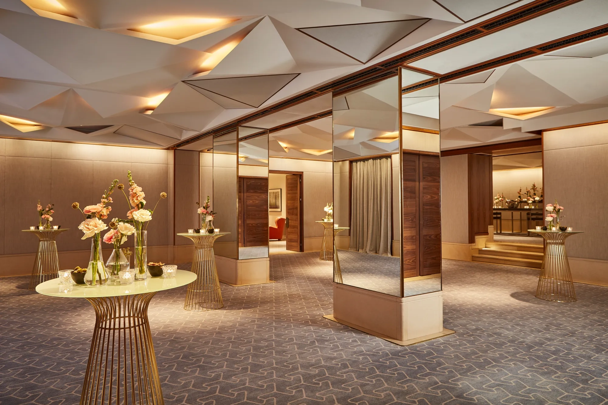 Elegant pre-function space with mirrored columns, gold tables, and floral decorations under geometric ceiling lights.