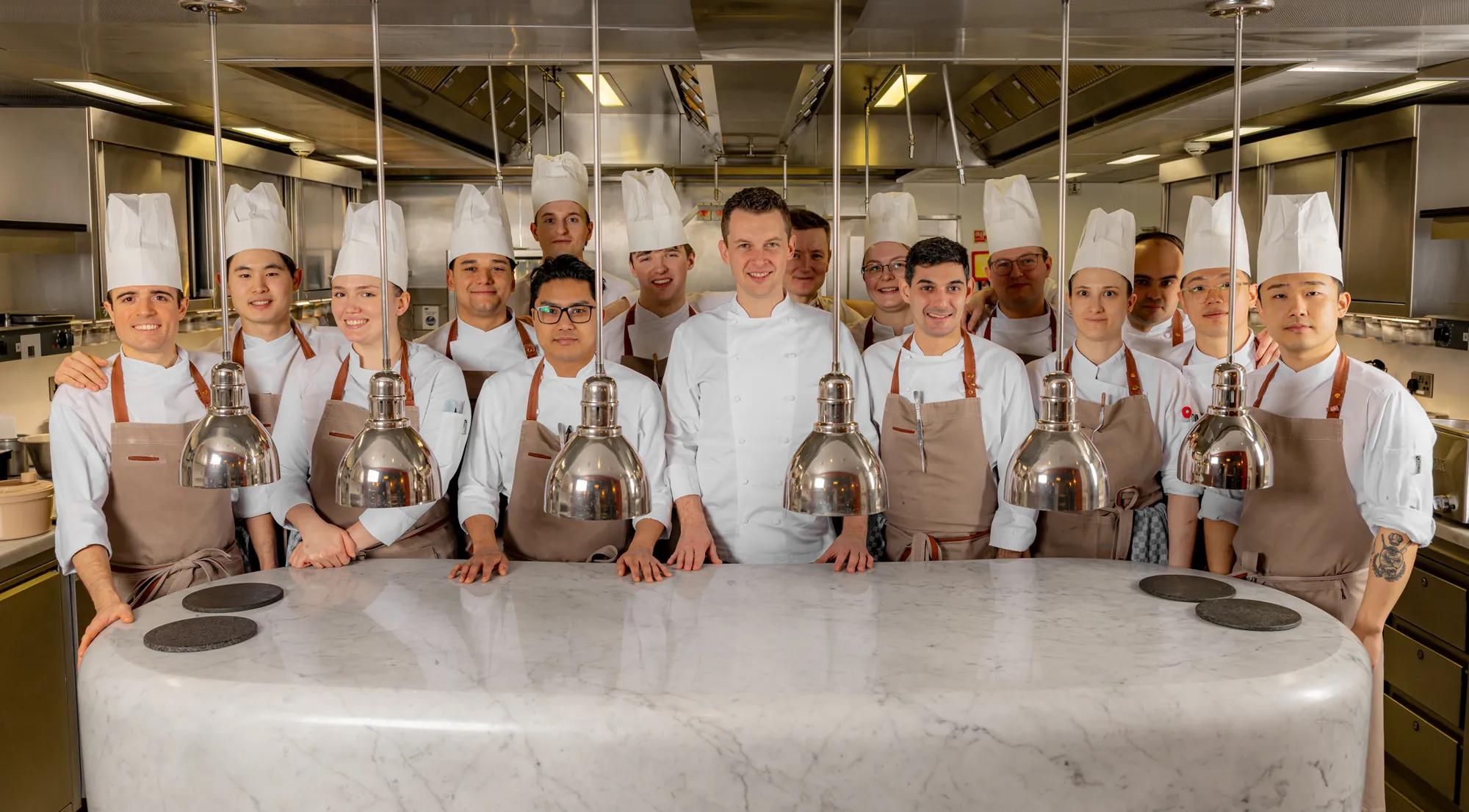 Group of chefs in white uniforms and aprons standing behind a marble pass in a professional kitchen