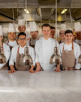 Group of chefs in white uniforms and aprons standing behind a marble pass in a professional kitchen