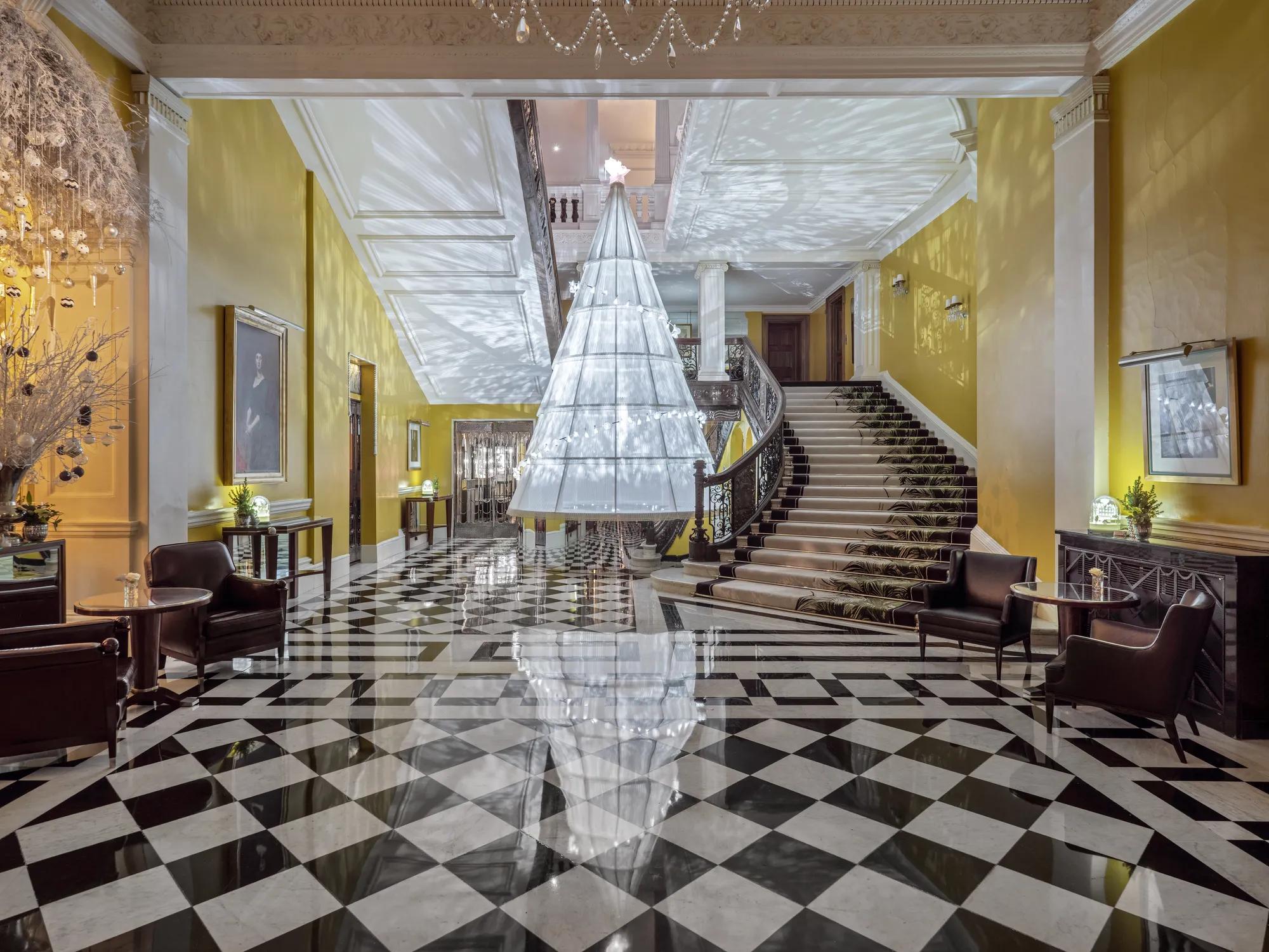 Grand hotel lobby with checkered marble floor and sweeping staircase, featuring a large suspended tree installation casting patterned light across yellow walls.