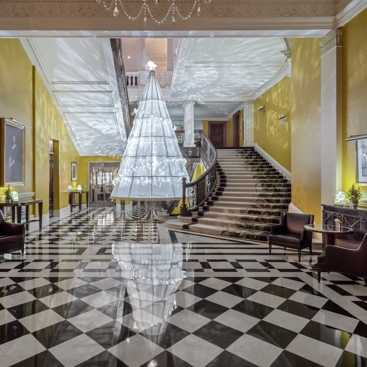Grand hotel lobby with checkered marble floor and sweeping staircase, featuring a large suspended tree installation casting patterned light across yellow walls.