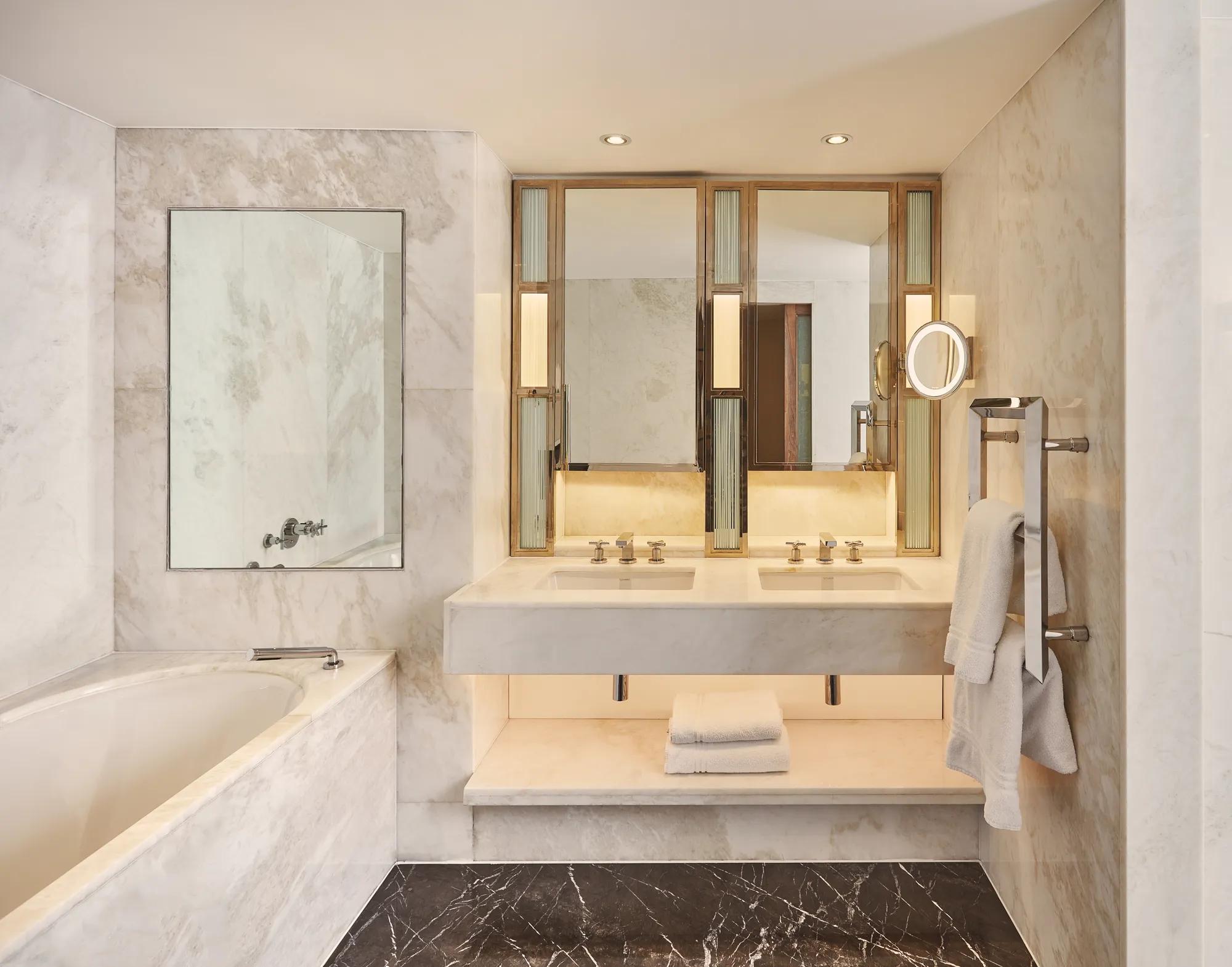 Marble bathroom with a bathtub, double vanity, illuminated mirrors, towel rack, and folded white towels on a shelf beneath the sink.